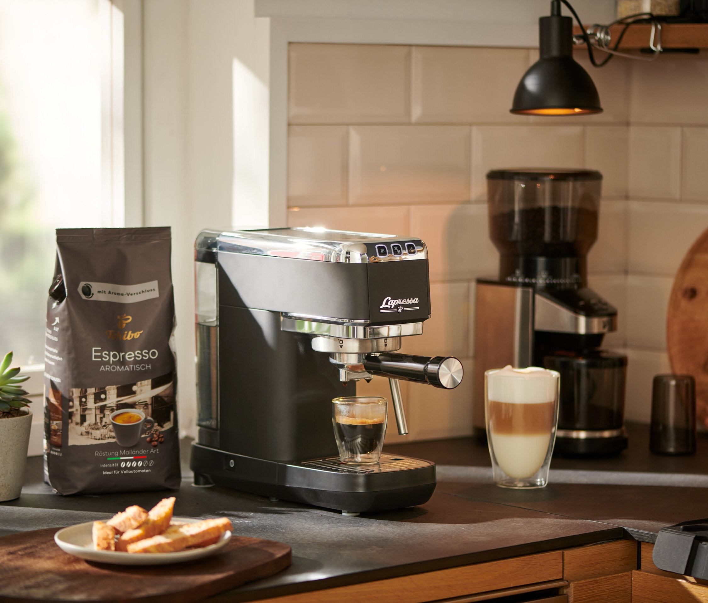 Eszpresszó folyik egy fekete »Lapressa« karos eszpresszó kávéfőzőből egy pohárba. A háttérben egy kávédaráló és egy csomag kávé látható.