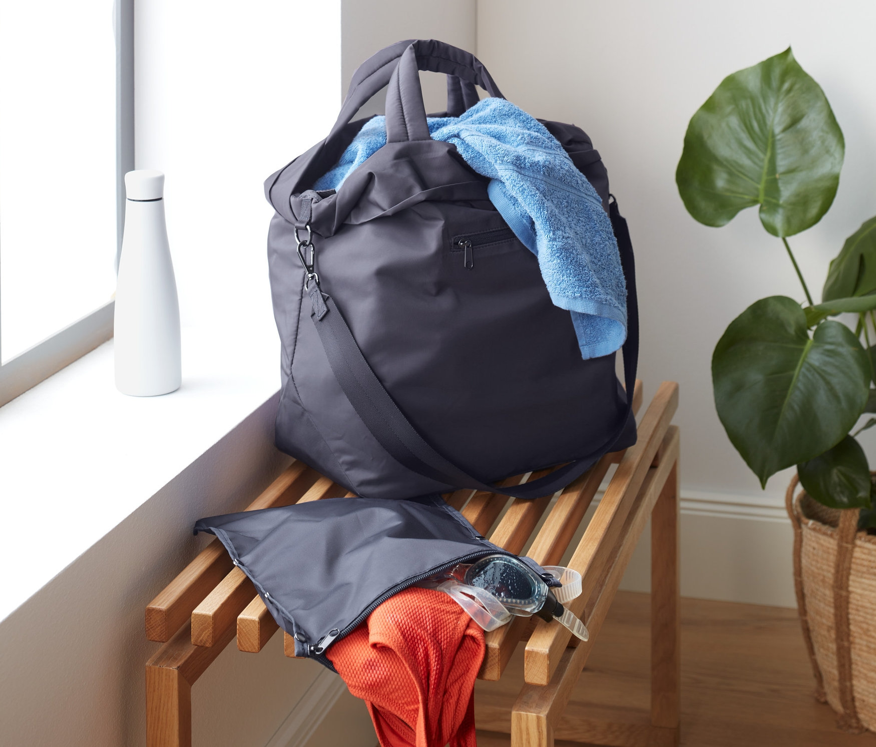 Egy sporttáska fekszik egy fa padon az ablak mellett. Egy kék törölköző lóg ki a táskából.