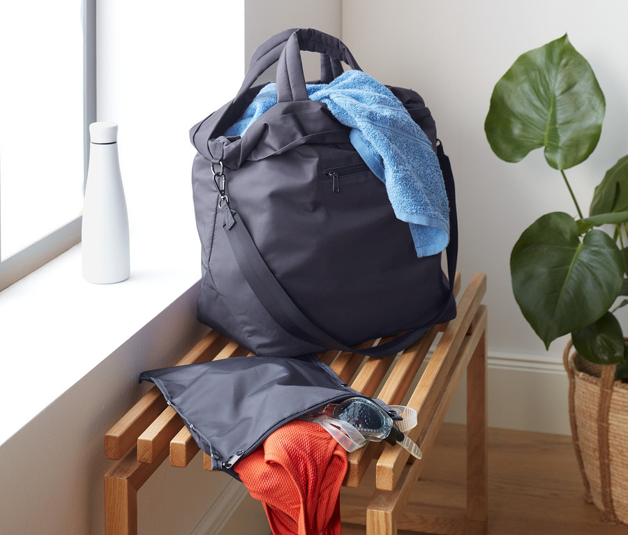 Egy sporttáska fekszik egy fa padon az ablak mellett. Egy kék törölköző lóg ki a táskából.