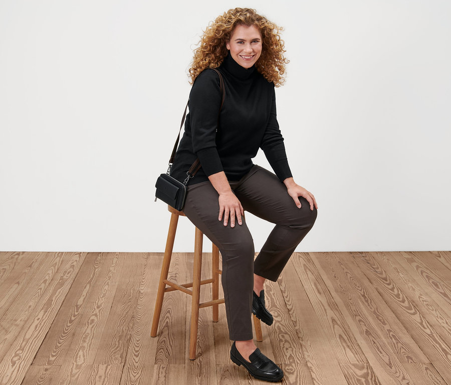 Nő ül a széken, fekete finom kötésű garbót, barna bengalin nadrágot, fekete bőr mokaszint és mini oldaltáskát visel.