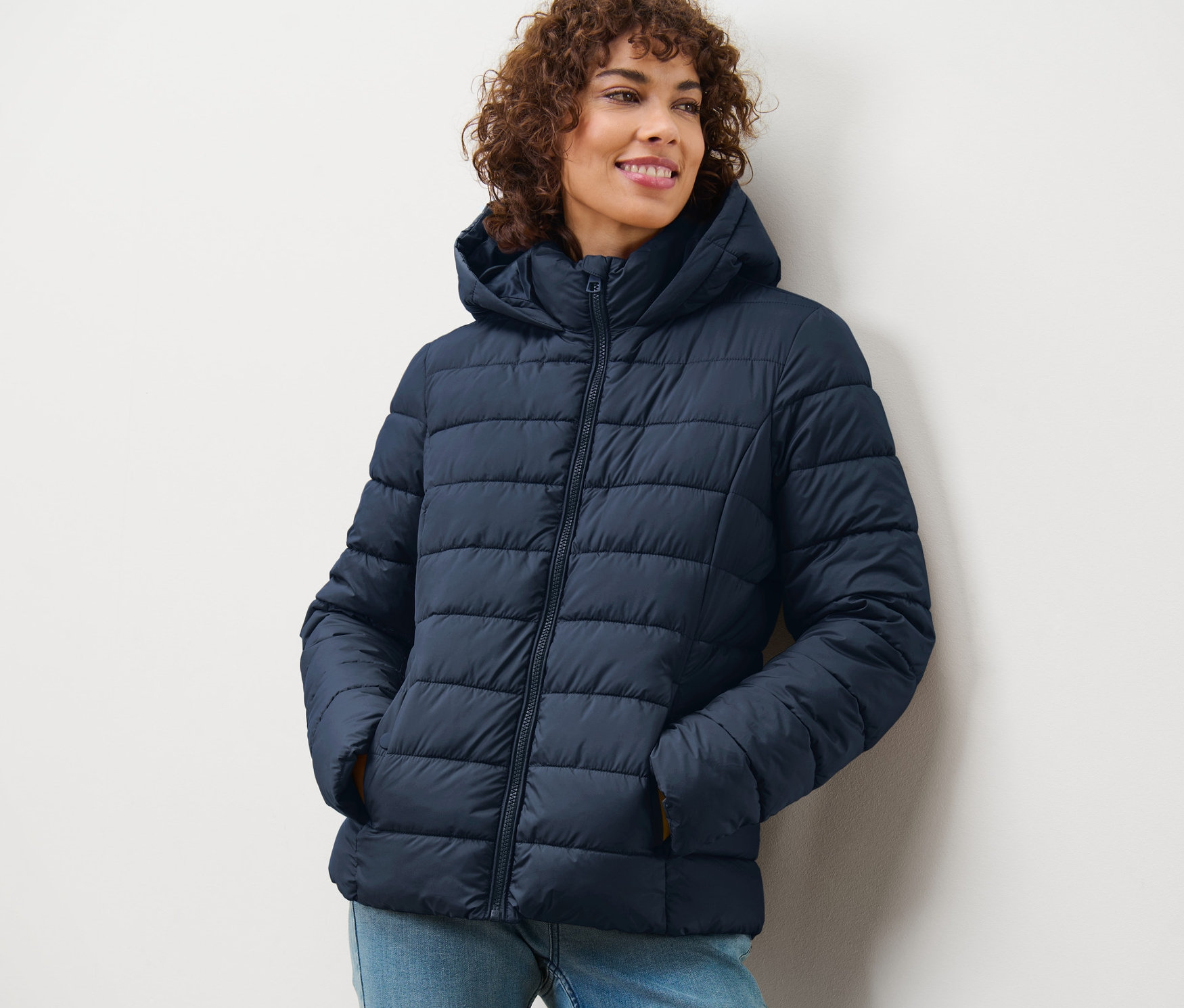 Nő mosolyog, sötétkék, steppelt, kapucnis dzsekit visel.