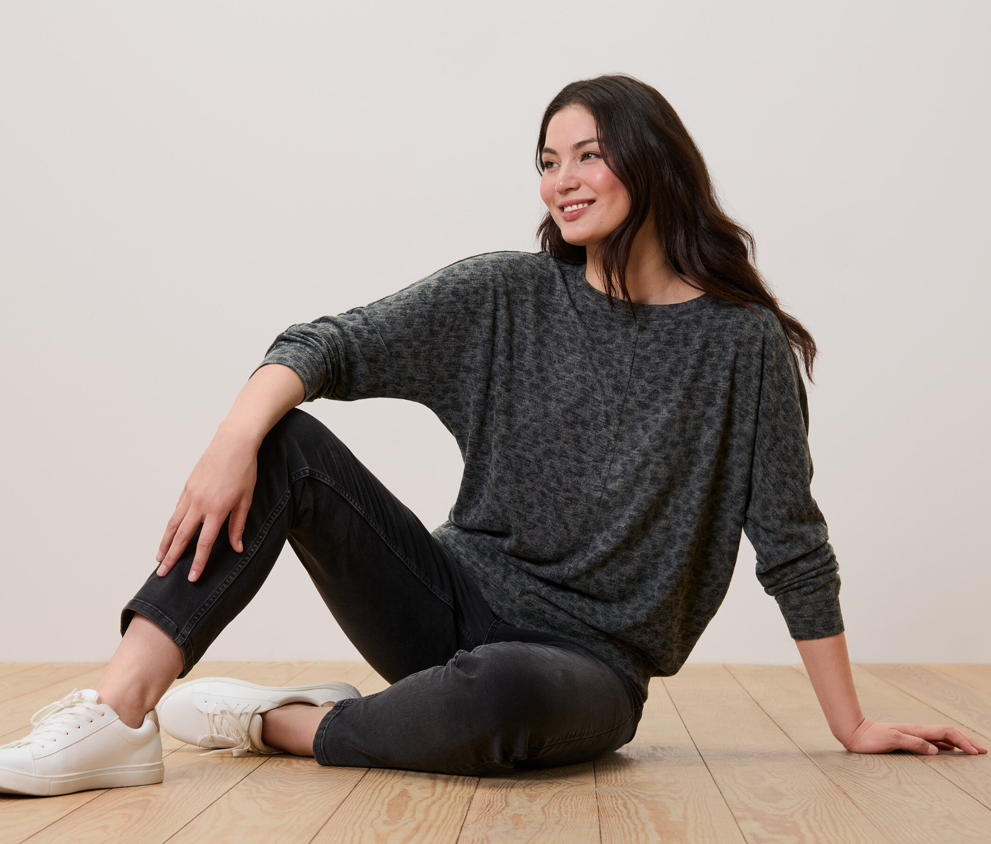 Nő ül a padlón, leopárdmintás denevérujjú felsőt, fekete farmert és fehér tornacipőt visel.