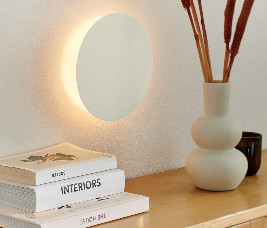 A falon egy LED-es fali lámpa, akkus. Egy fa komódon váza díszágakkal és könyvek.