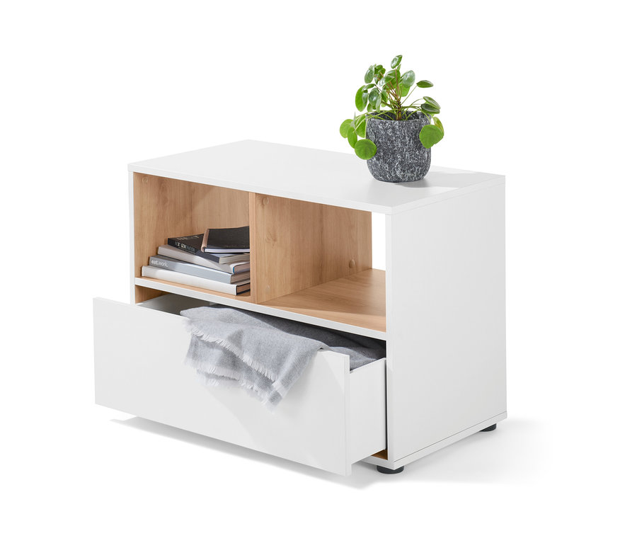 Fehér komód fiókkal, tetején egy növény szürke cserépben. A nyitott rekeszben könyvek, a fiókban szürke takaró található.