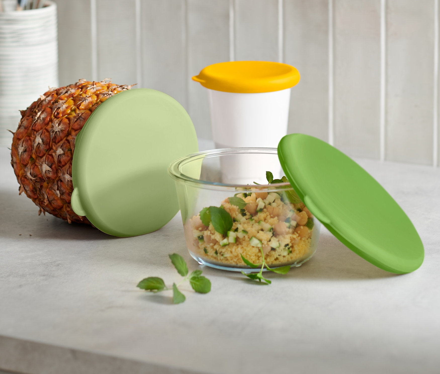 Ananász a 3 szilikon frissentartó fedél egyikével és üvegtál a második zöld fedéllel egy sárga fedővel ellátott fehér edény mellett.