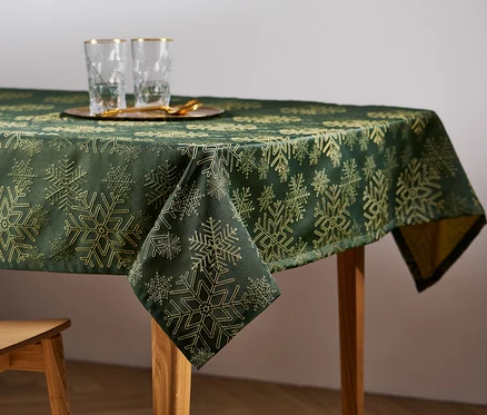 Egy fa asztalon zöld, hópelyhes mintázatú jacquard asztalterítő látható. Az asztalon poharak és egy fa szék áll.
