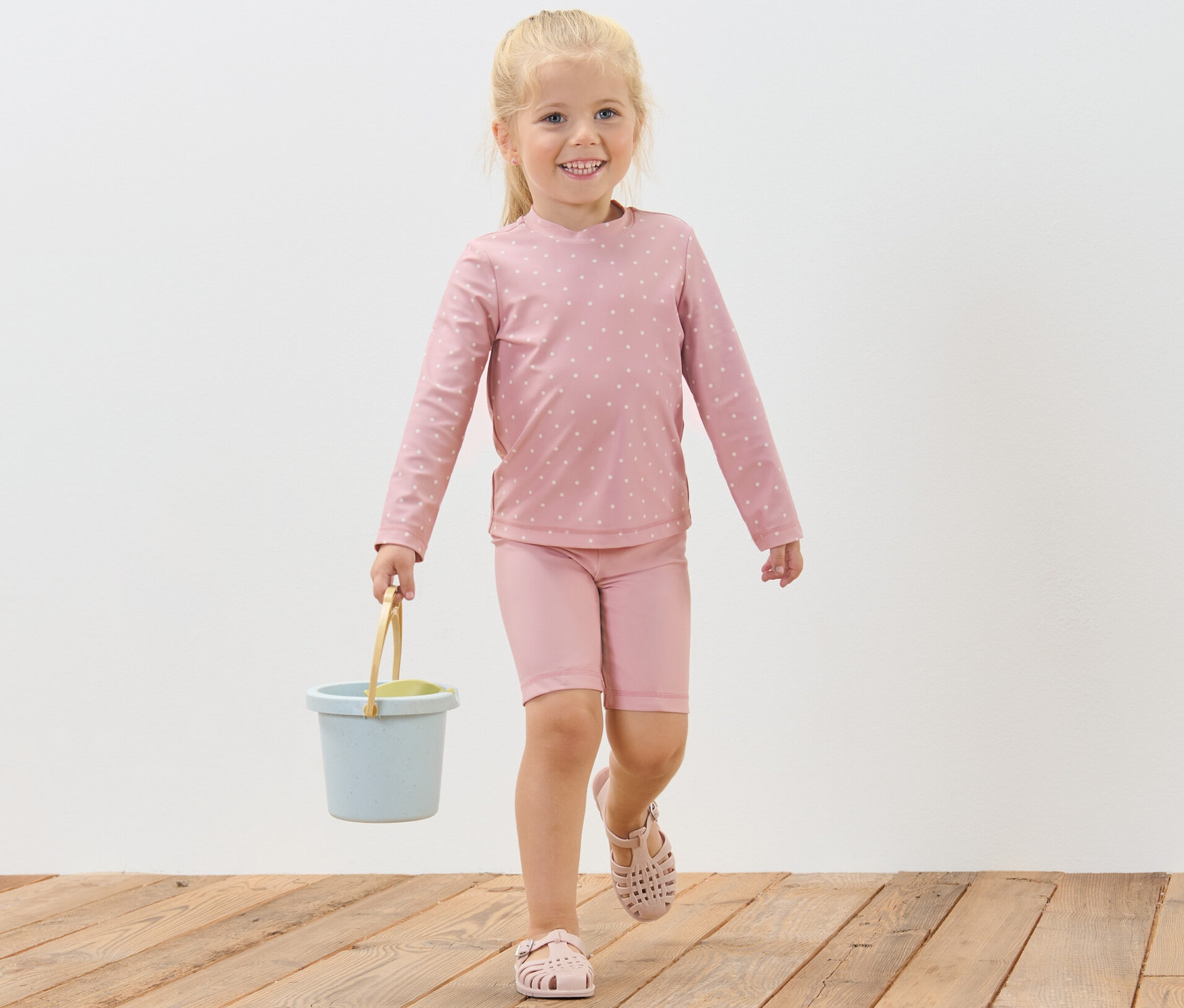 Egy mosolygós kislány sétál egy fa padlón, és egy kék vödröt visz. Kisgyerek UV-szűrős együttest visel, pöttyös.