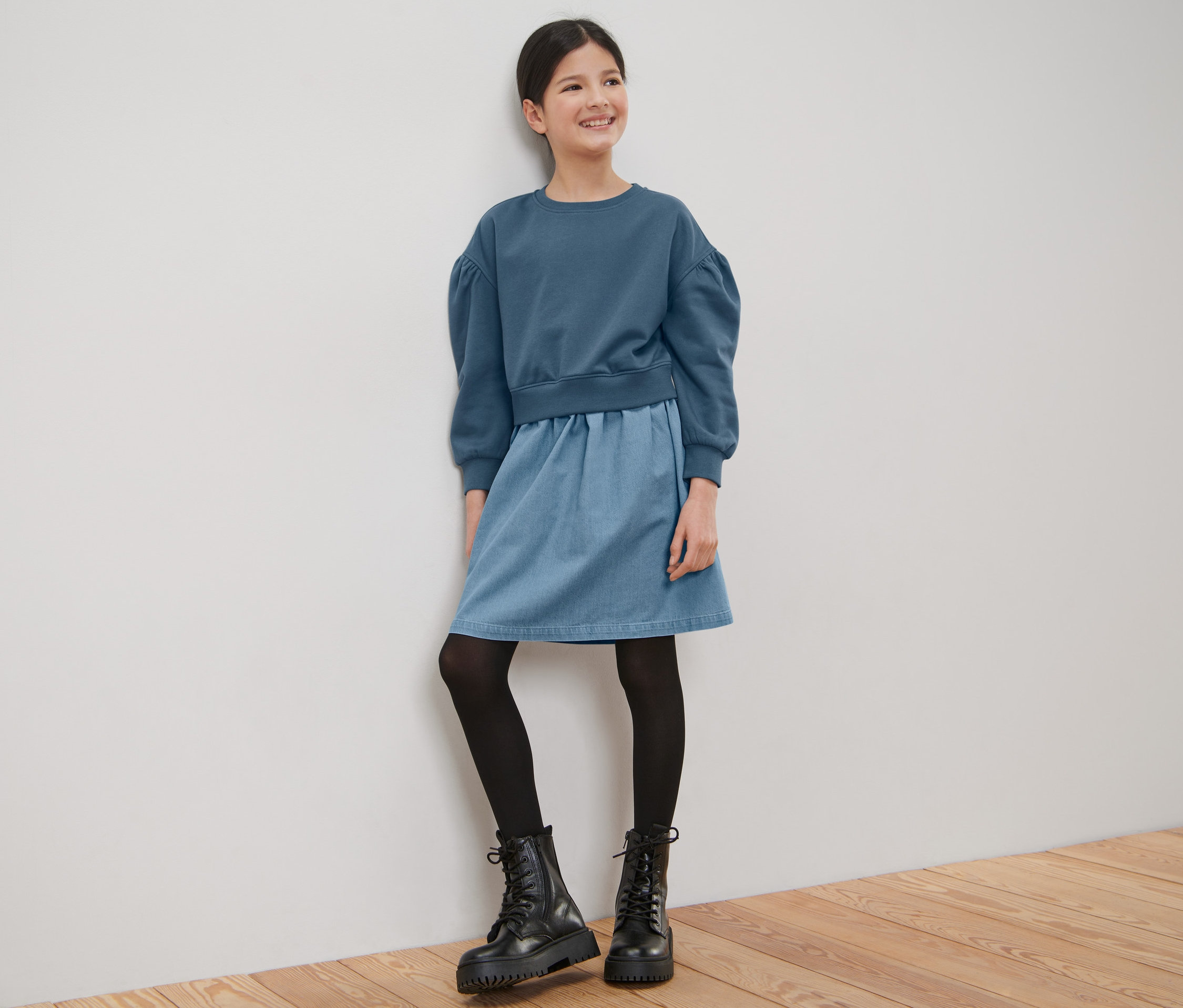 Mosolygó lány lányka kék szabadidőruhában a falnak támaszkodva áll.