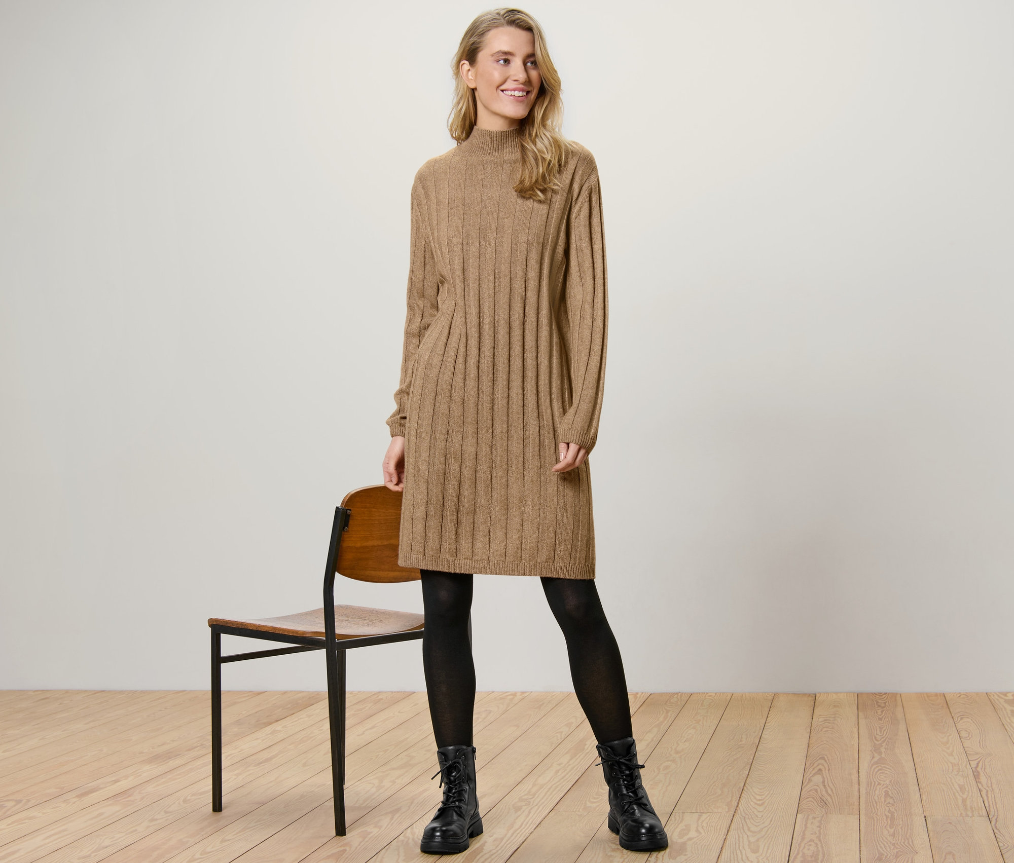 Egy nő áll egy szék mellett, és egy világosbarna kötött ruhát visel.