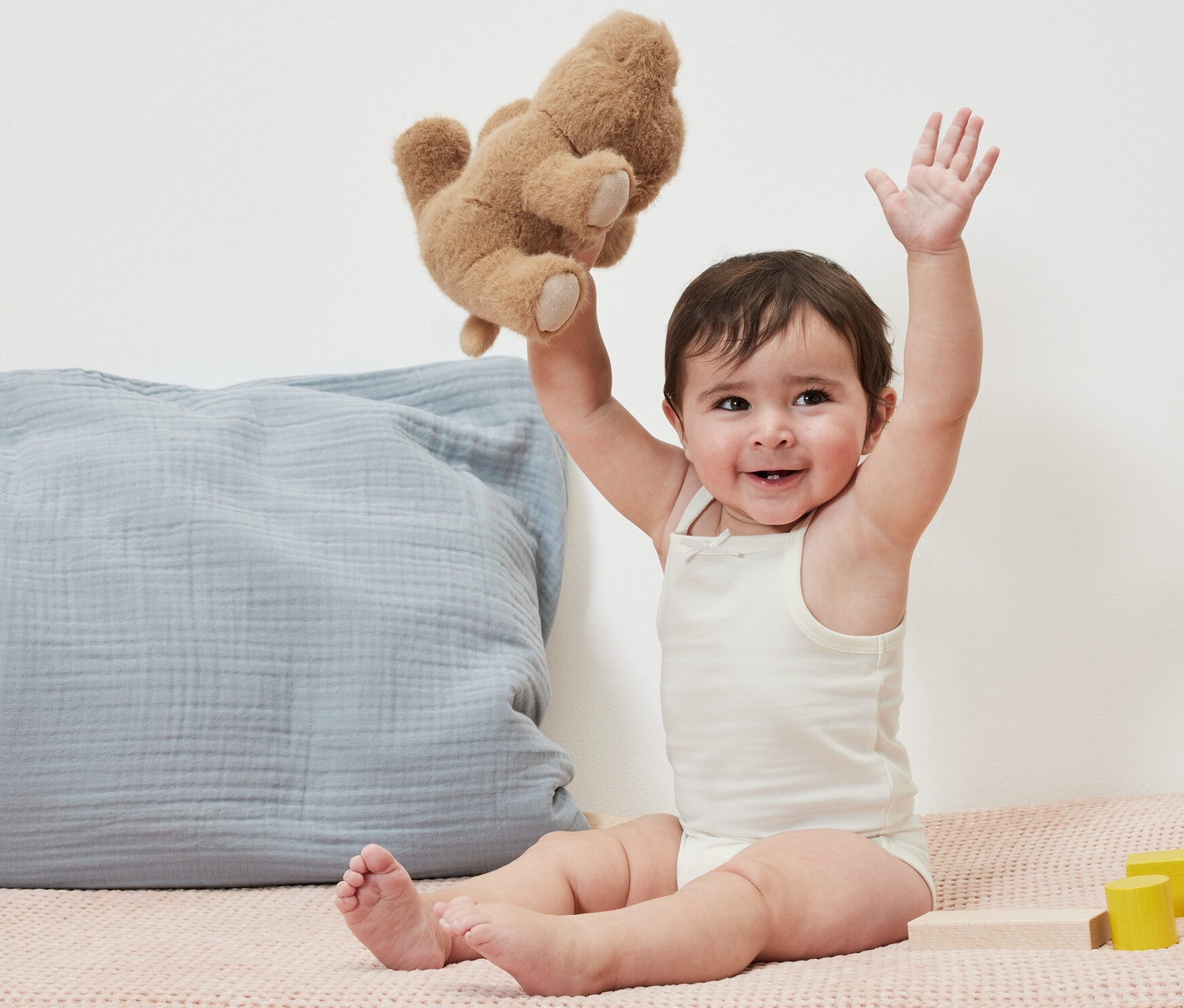 Egy mosolygó baba ül és egy plüssmackót tart a kezében. Rajta 5 kisgyerek body.