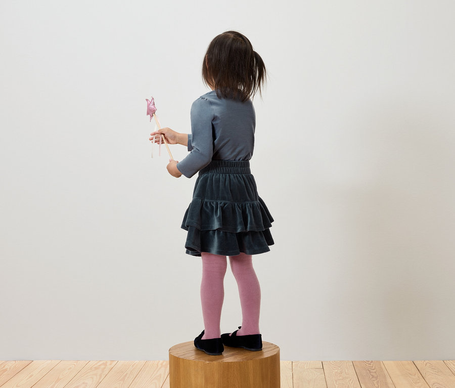 Szürke pólót és zöld szoknyát viselő lány áll egy fa talapzaton, és egy pálcát tart.