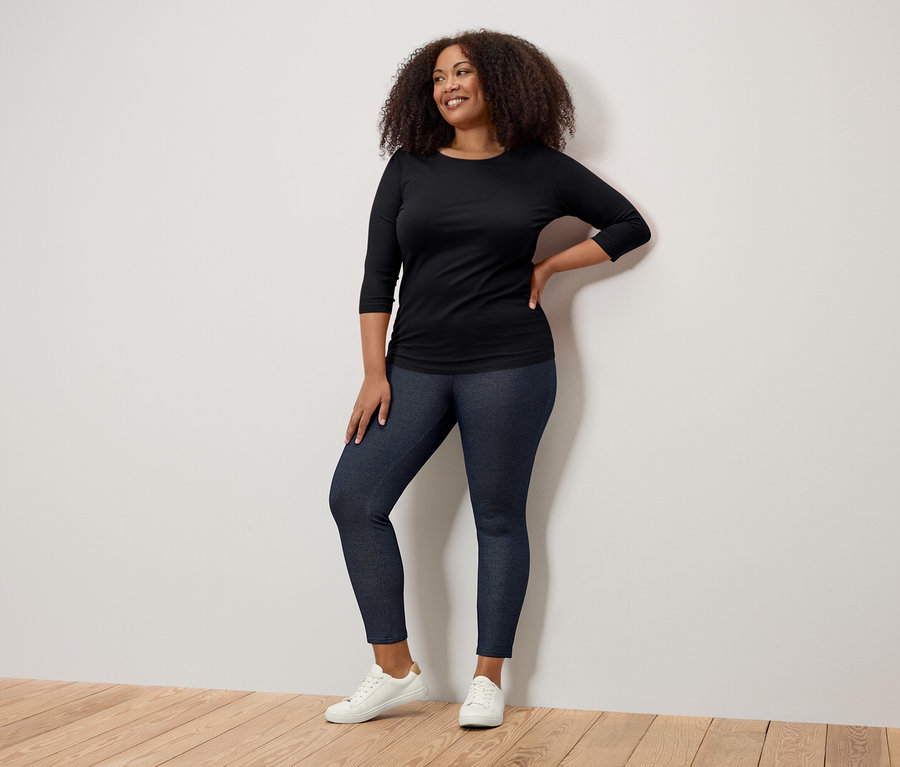 Mosolygó nő pózol fekete háromnegyedes ujjú pólóban, sötétkék jeggingsben és fehér tornacipőben.