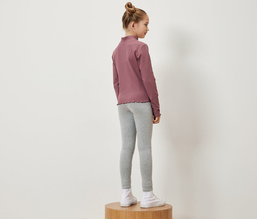 Egy lány oldalra áll egy fa talapzaton, rózsaszín hosszú ujjú inget, szürke leggingset és fehér tornacipőt visel.