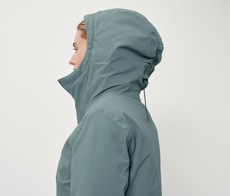 Egy nő profilja kékeszöld kabátban, kapucnival, fehér háttér előtt.