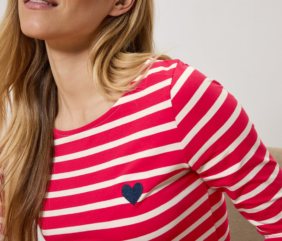 Egy szőke nő piros-fehér csíkos, hosszú ujjú inget visel sötétkék szívvel.