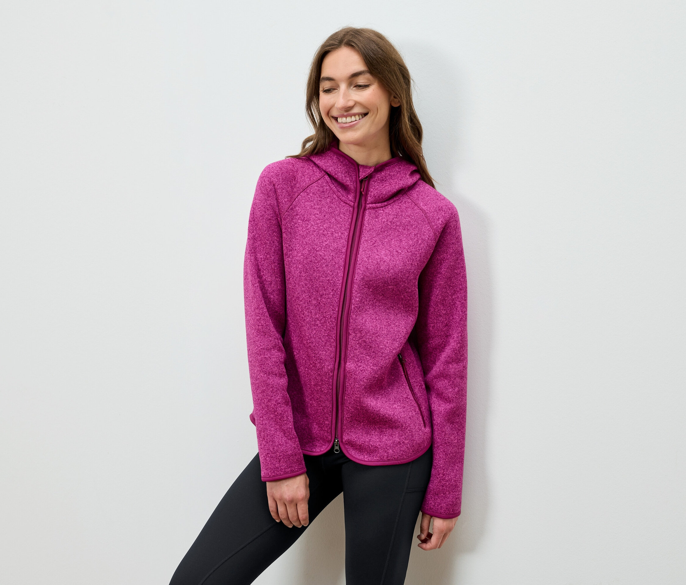 Mosolygó nő pózol rózsaszín kapucnis pulóverben és fekete nadrágban.