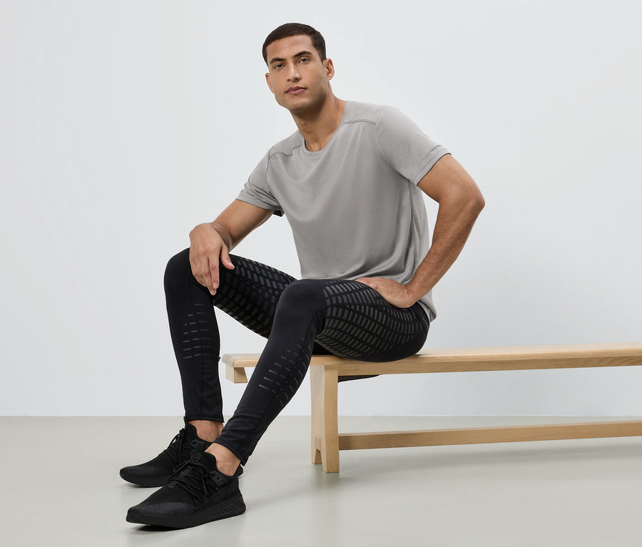 Férfi ül egy padon, szürke pólót, mintás fekete leggingset és fekete tornacipőt visel.