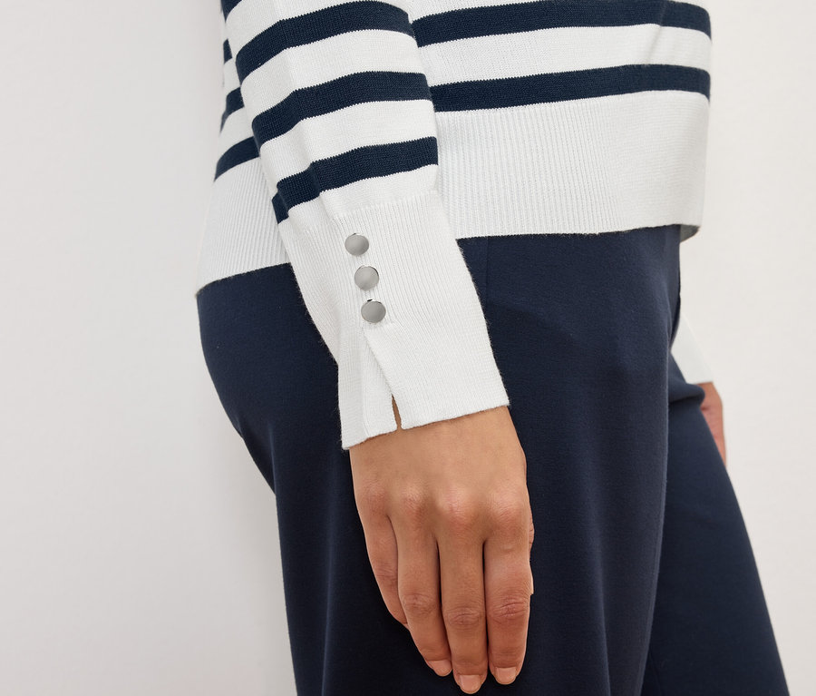 Egy nő csíkos, hosszú ujjú pulóvert visel, gombokkal a mandzsettán és sötétkék nadrágot.