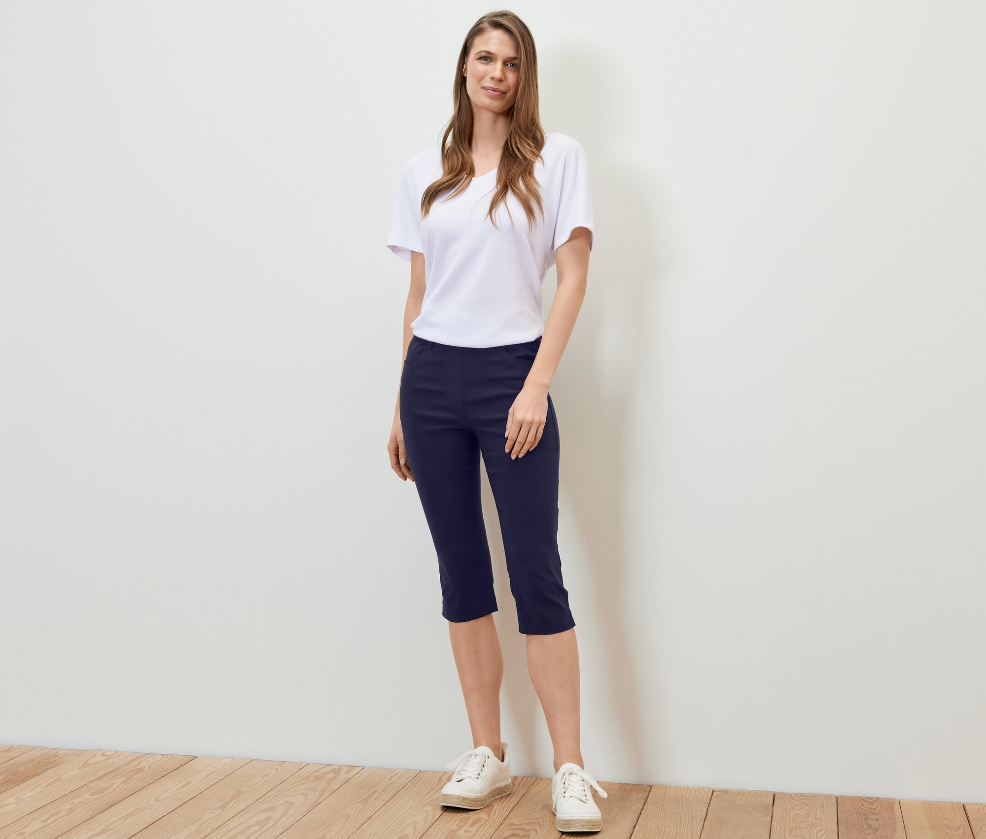 Egy nő áll, sötétkék 3/4-es bengalin nadrágot, fehér pólót és tornacipőt visel.