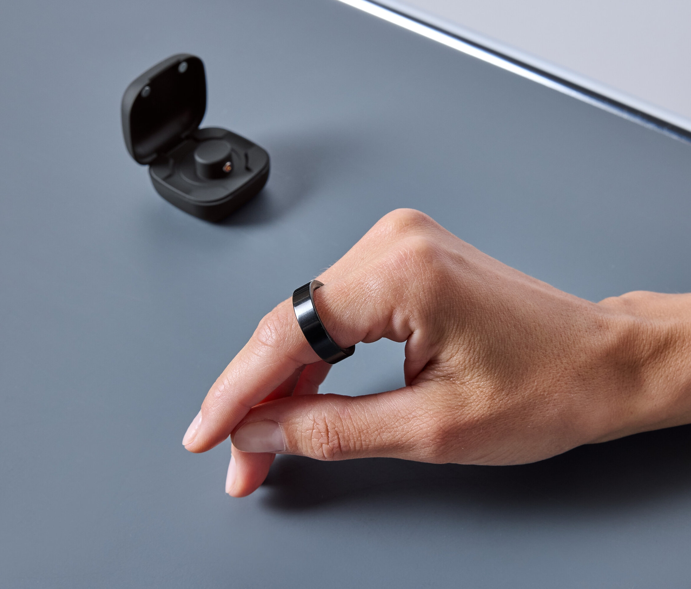 Egy kéz fekete gyűrűt visel. A gyűrű mellett egy nyitott tok található.