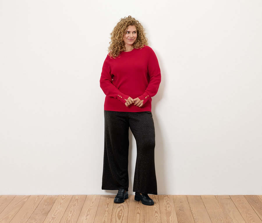 Göndör hajú nő áll, piros pulóvert és fekete nadrágot visel.