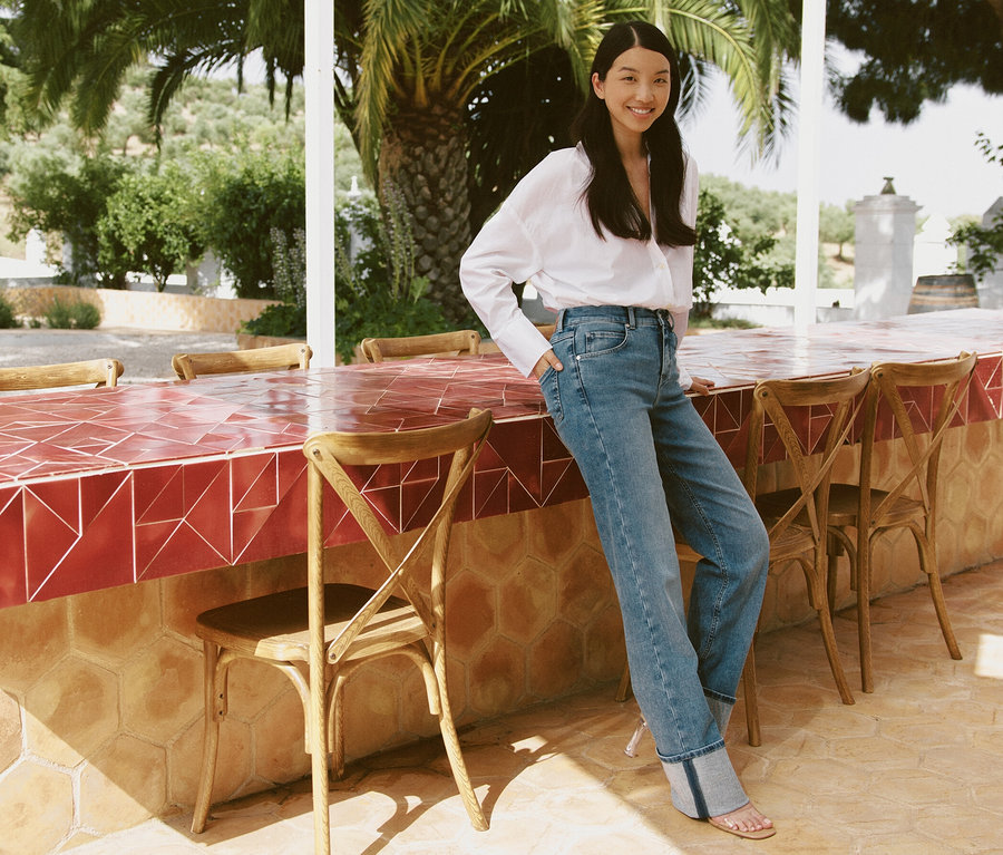 Egy nő egy kültéri bárpultnak dőlve pózol, világoskék Angels »Lara« Straight Fit feltűrt szárú farmerben.