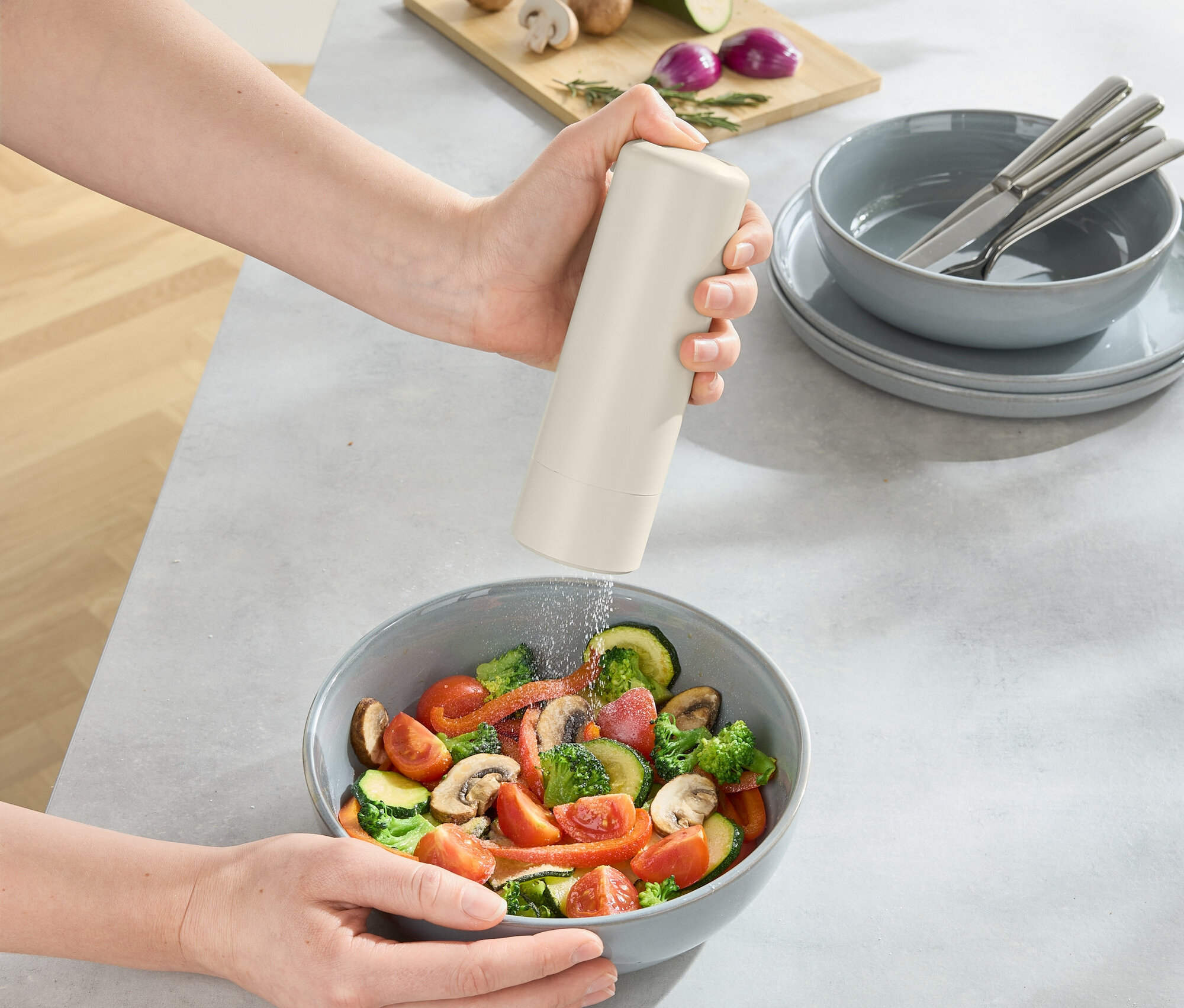 Egy személy fűszert szór egy salátástálba, az asztalon pedig egy deszka gombával, hagymával és cukkinivel.