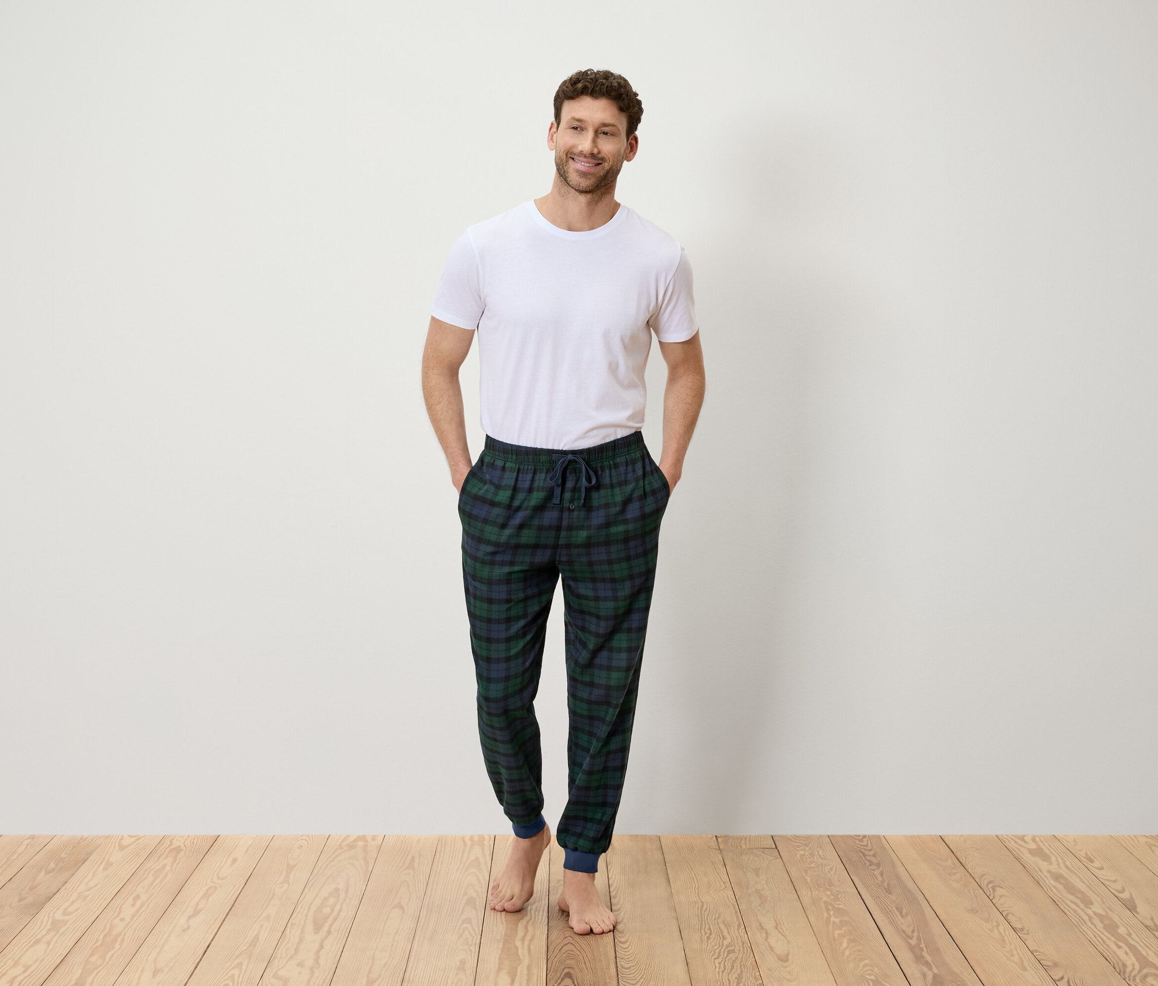 Egy férfi áll zsebre tett kézzel, fehér pólót és zöld-kék kockás nadrágot visel.