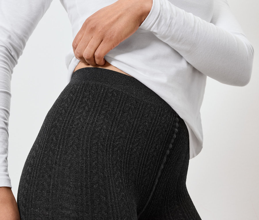 Egy nő szürke kötött leggingsben és fehér hosszú ujjú felsőben igazítja a leggings derekát.