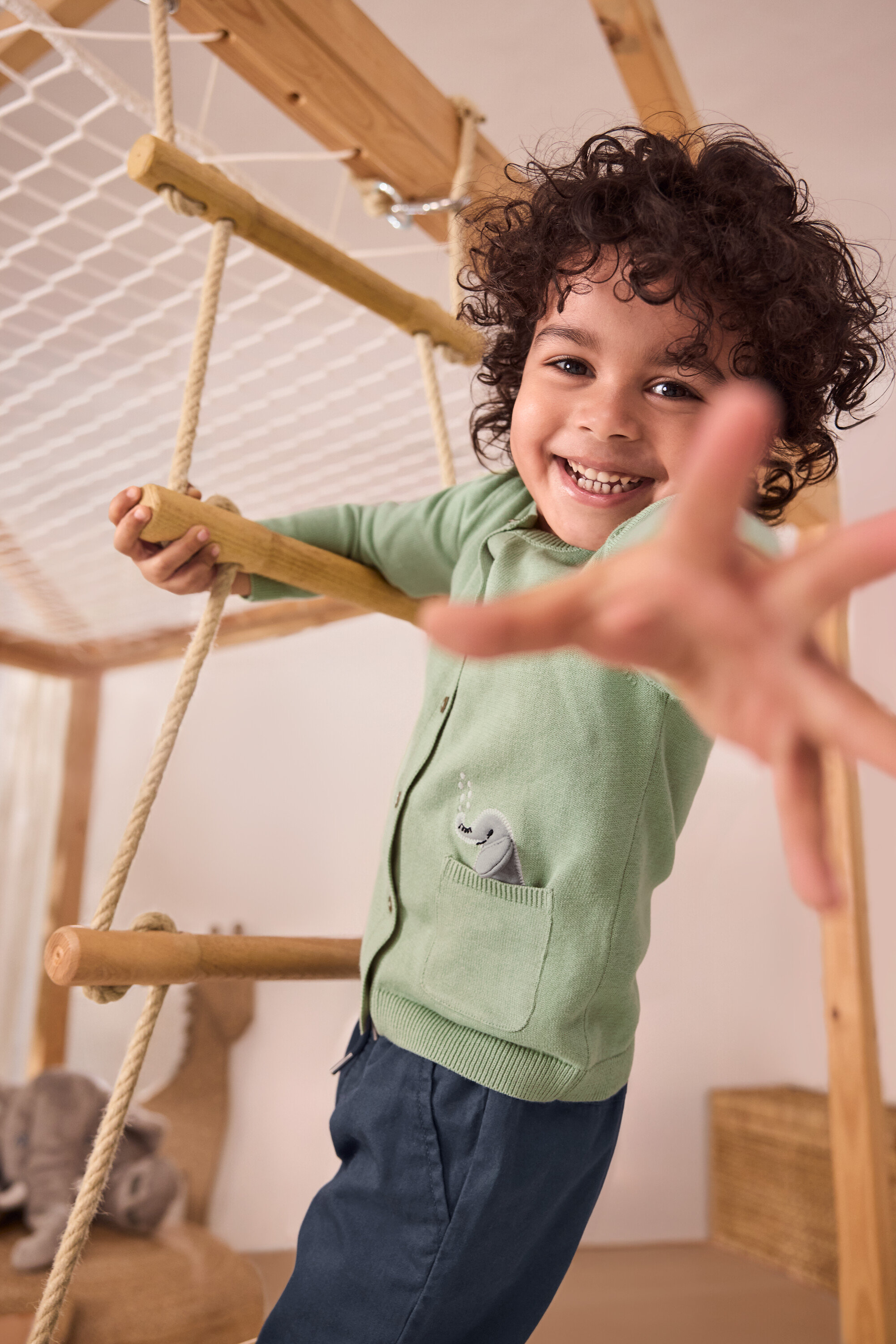 Egy mosolygós fiú mászik egy kötél létrán. Zöld Kinder-Strickjacke mit Applikation és kék Belebújós nadrág gyerekeknek – Fit »Anton« van rajta.