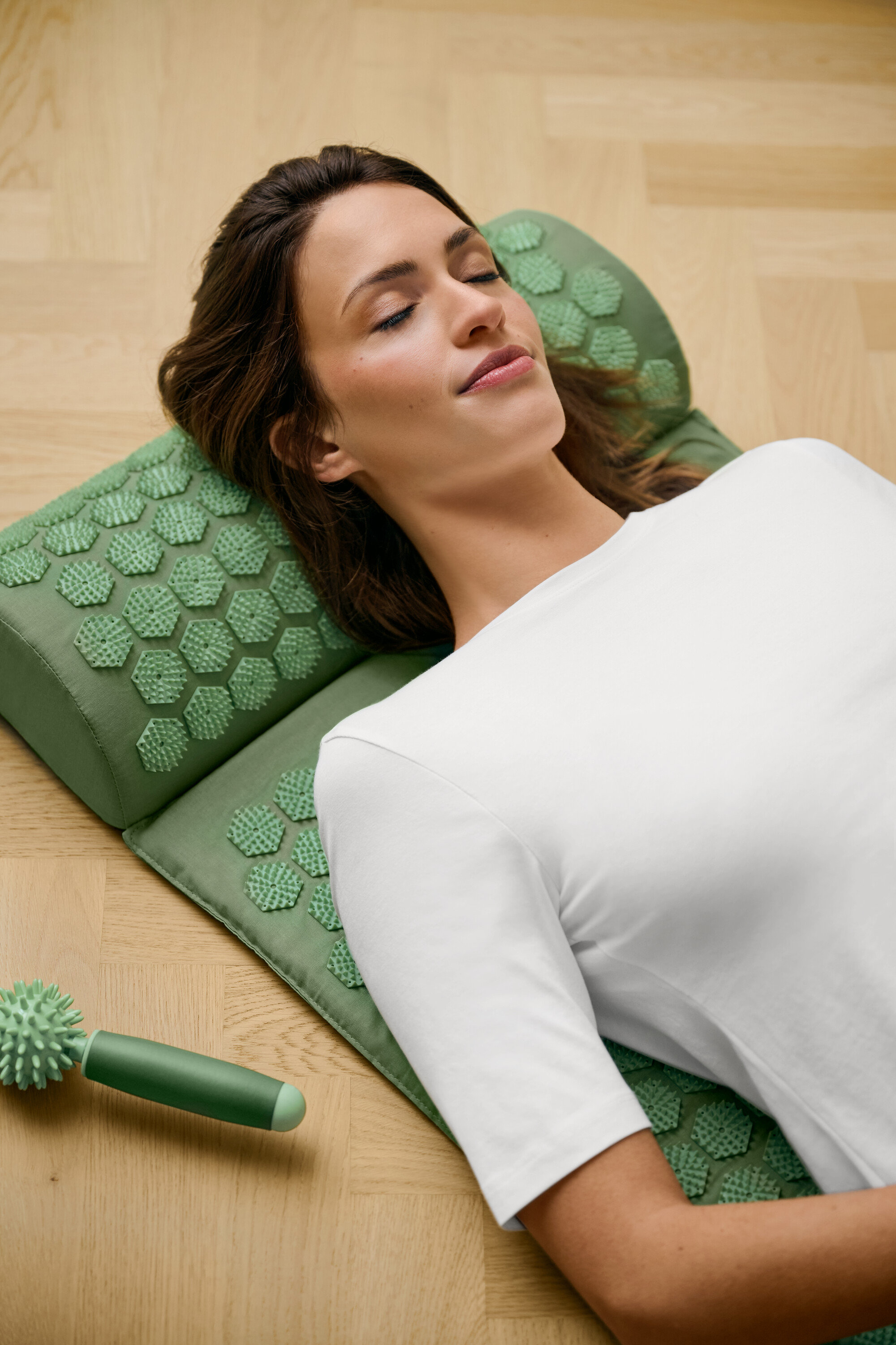 Nő fekszik egy zöld akupresszúrás szőnyegen, és fehér, félhosszú ujjú felsőt visel.