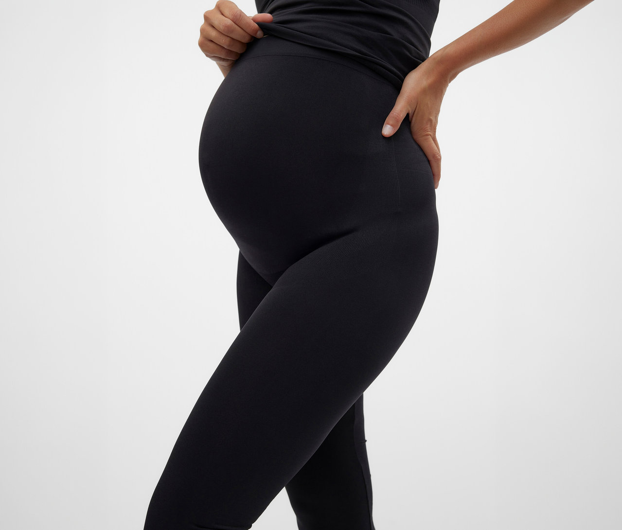 Fekete felsőt és MAMALICIOUS kismama leggingset viselő terhes nő pózol fehér háttér előtt.