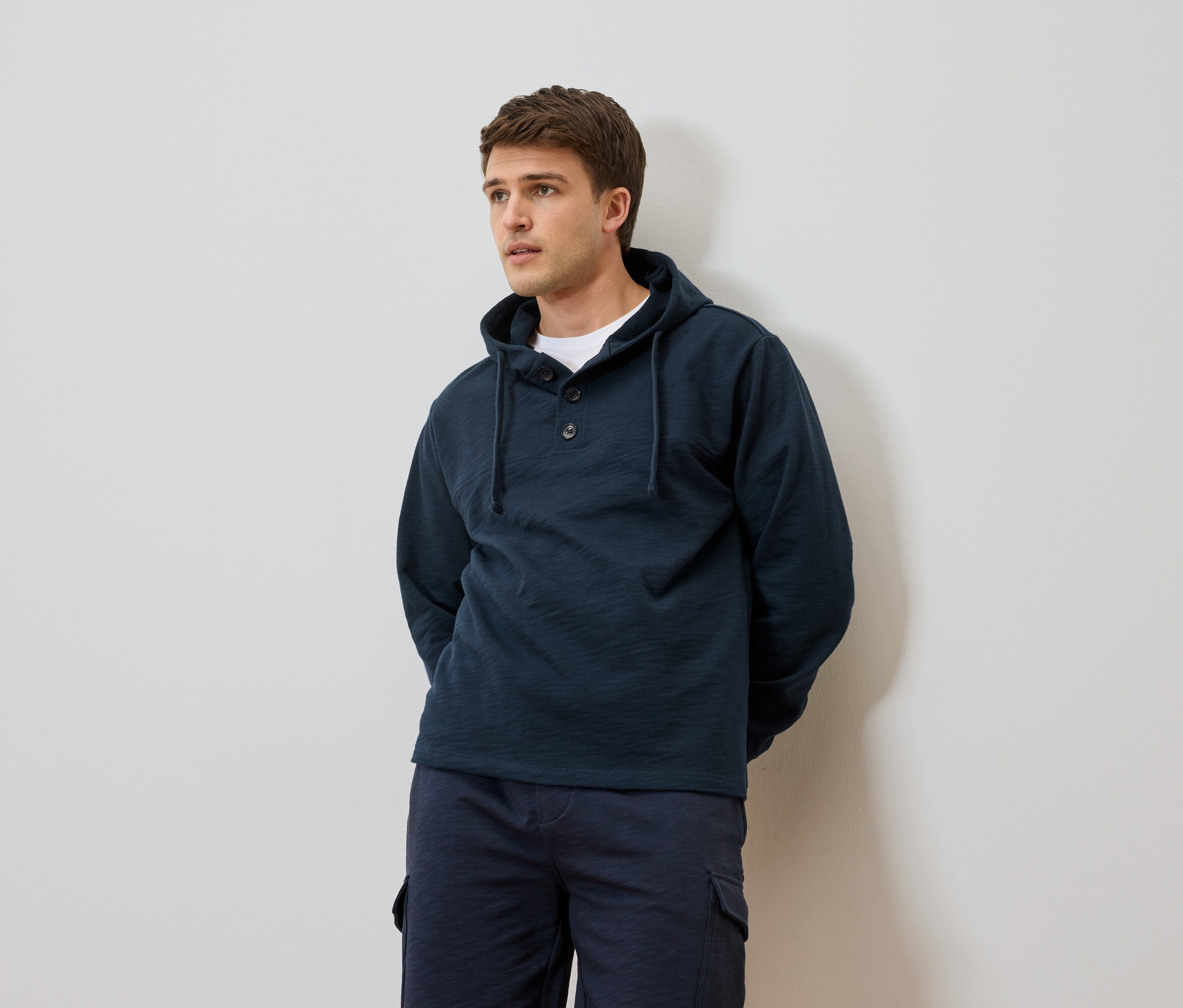 Férfi sötétkék kapucnis pulóverben és rövidnadrágban, fehér fal előtt.