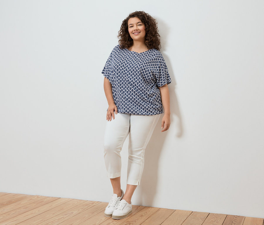 Göndör hajú nő kék-fehér mintás blúzban, fehér nadrágban és fehér tornacipőben áll egy fehér fal előtt.