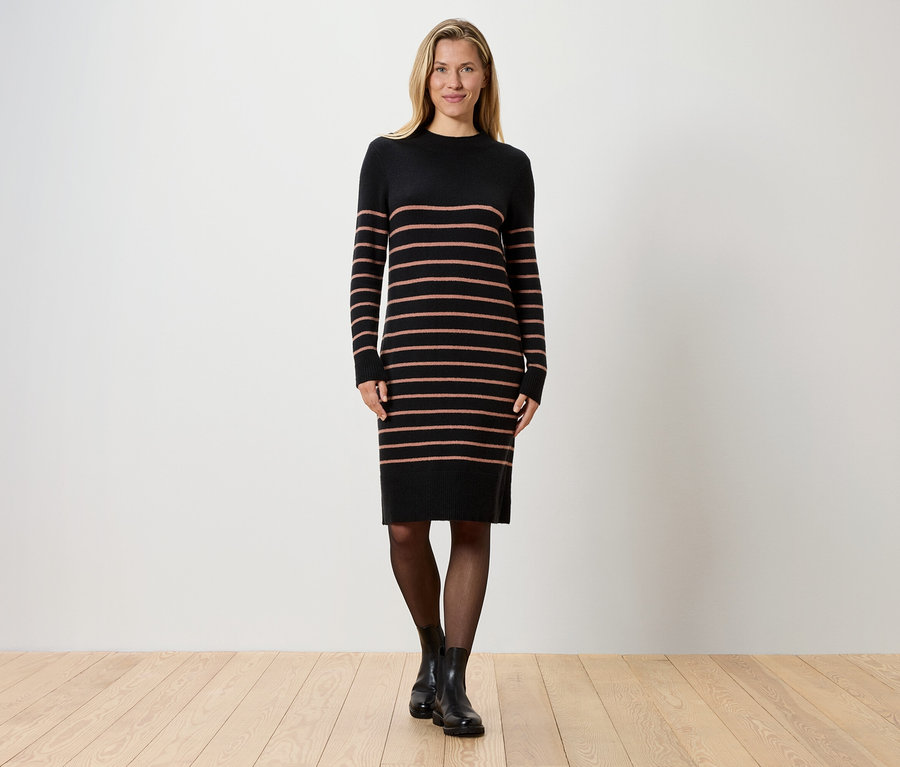 Egy szőke nő áll egy fehér fal előtt, csíkos, fekete kötött ruhát, fekete harisnyát és fekete csizmát visel.