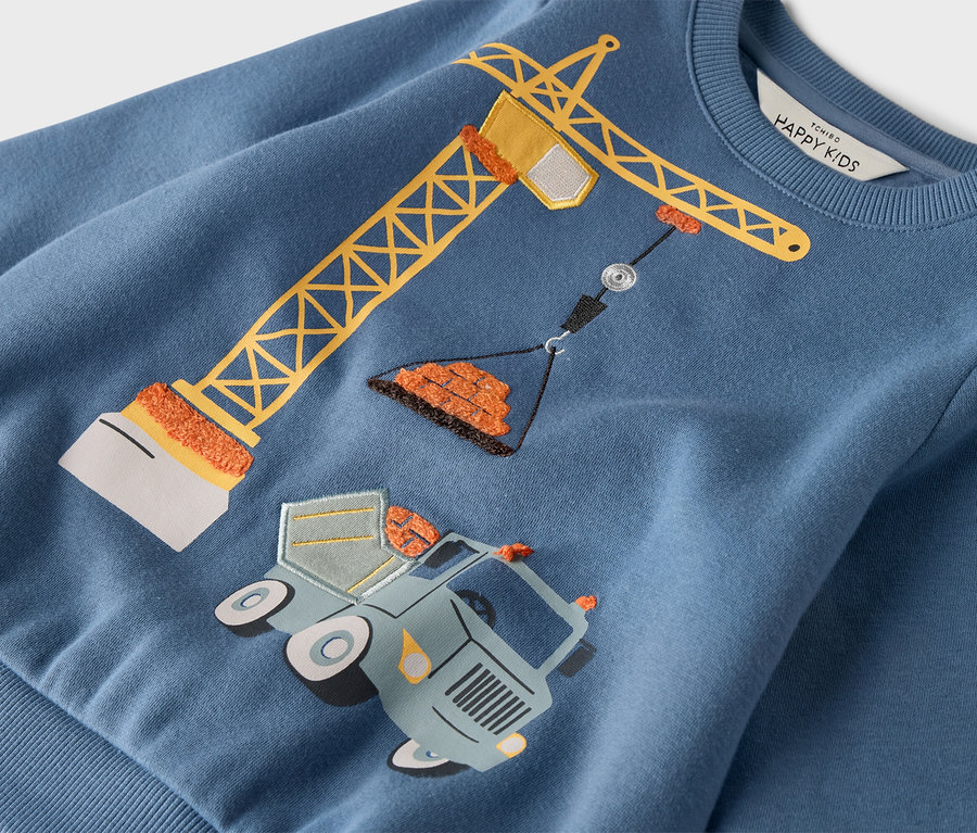 A kék pulóveren egy sárga daru látható, amely egy kerekes teherautó fölött áll.