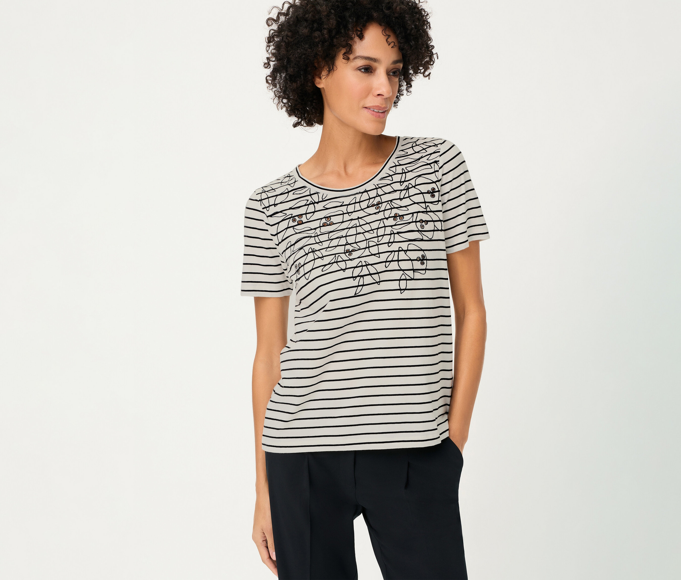 A göndör hajú nő virágmintás, csíkos Olsen pólót és sötét nadrágot visel.