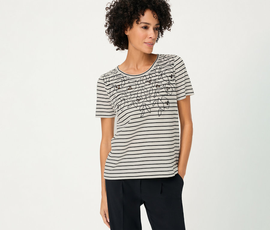 A göndör hajú nő virágmintás, csíkos Olsen pólót és sötét nadrágot visel.