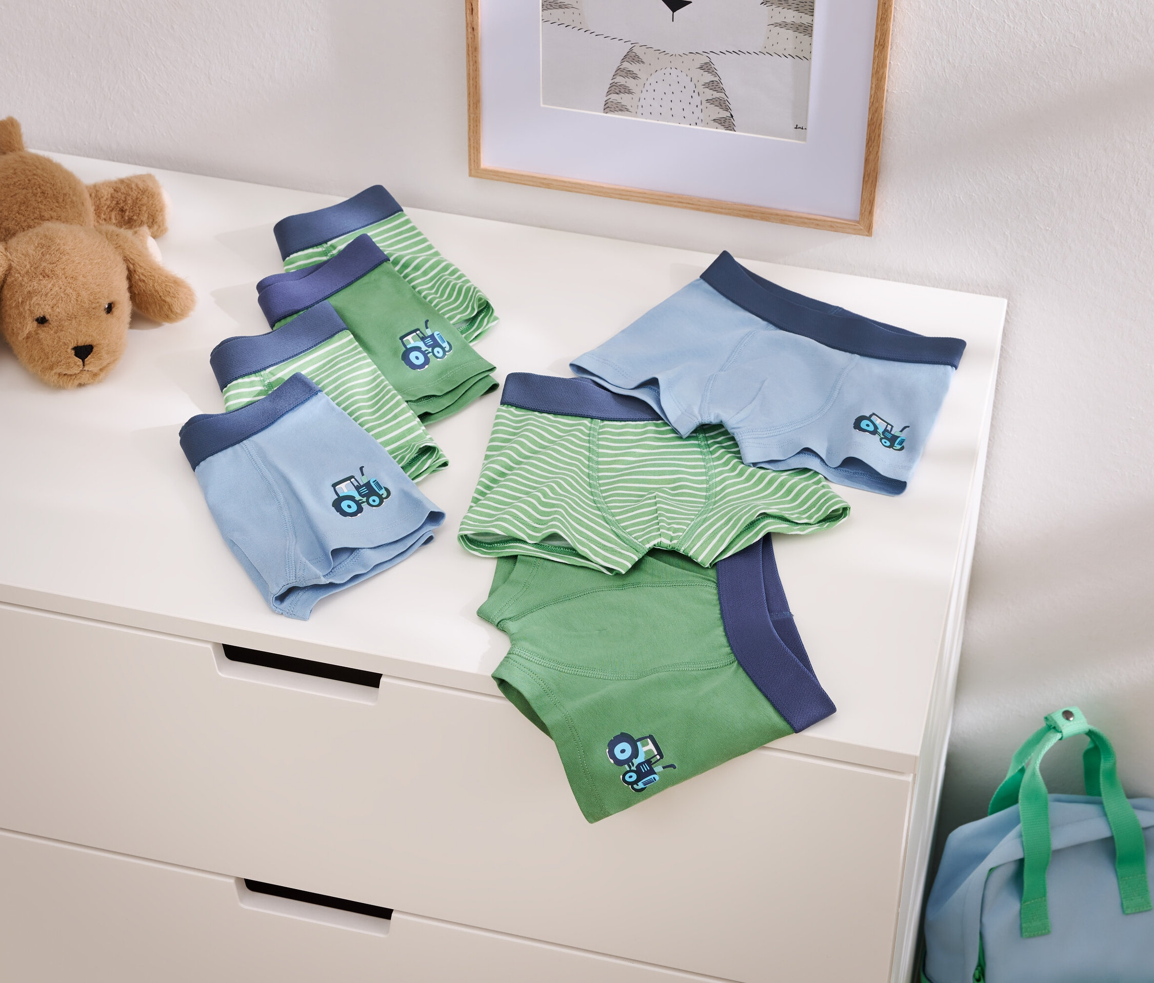 Fotó világoskék és zöld fiú boxeralsókról traktor mintával egy komódon, egy plüsskutya és egy kék utazótáska mellett.