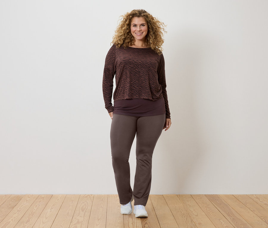 A göndör hajú nő barna mintás, hosszú ujjú felsőt, sötétebb színű topot és barna nadrágot, valamint fehér tornacipőt visel.