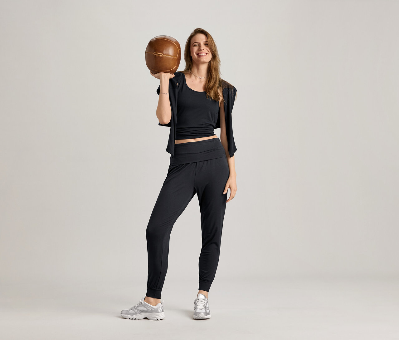 Nő fekete 2 az 1-ben sportfelsőben, tréningnadrágban és sportcipőben tart egy barna labdát.