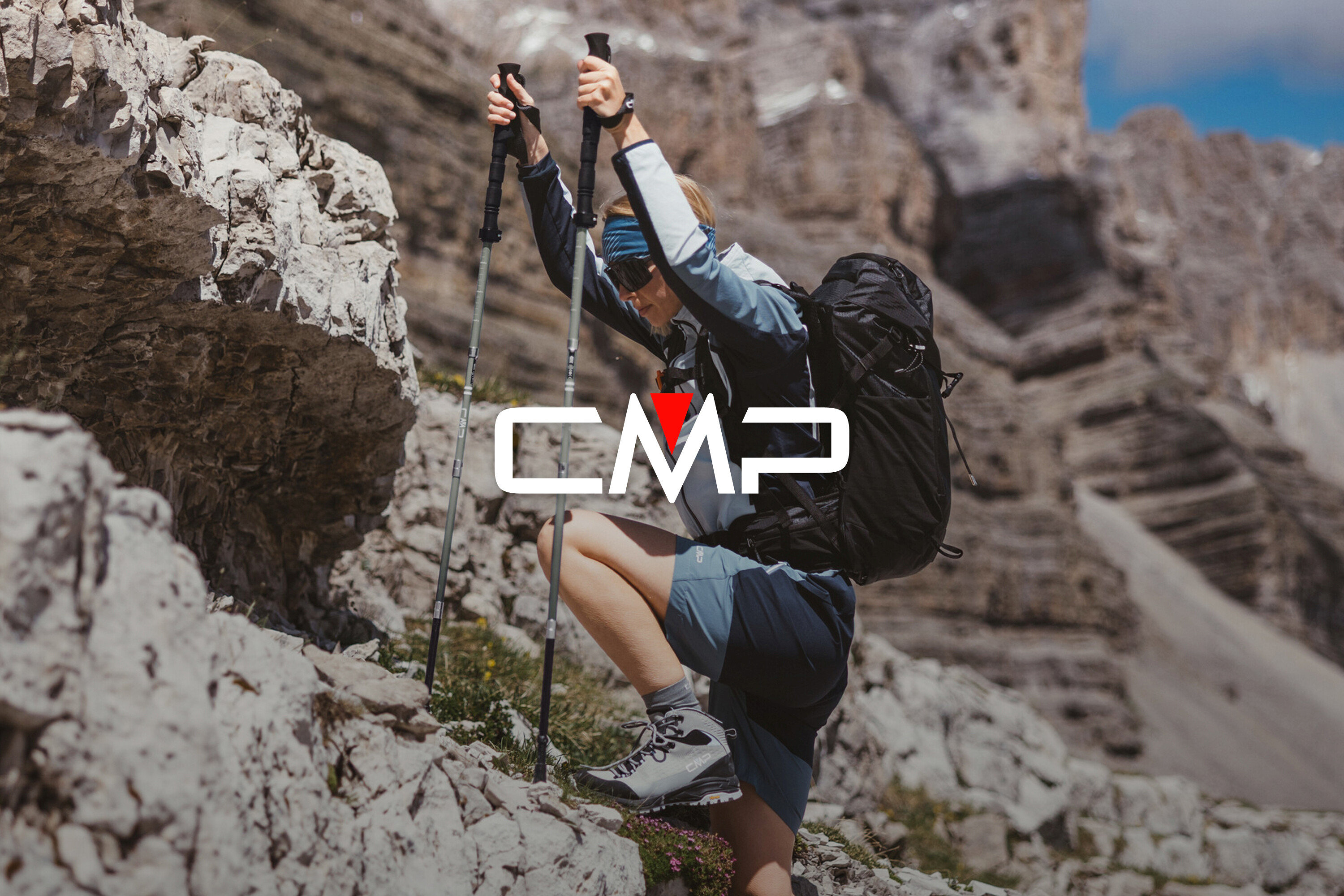 Egy hátizsákos, túrabotos nő sziklákat mászik. Túrabakancsot, rövidnadrágot és kabátot visel.