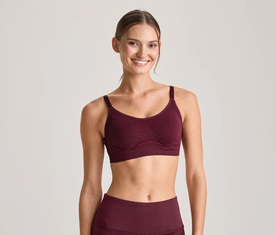 Mosolygó nő bordó sportmelltartóban és hozzá illő leggingsben, a kamerába néz.