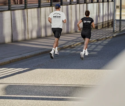 Egy férfi fehér sportfelsőben „RUN CLUB” felirattal, fekete sport rövidnadrágban és uniszex Coffe Crew teniszzokniban, valamint egy nő fekete sportfelsőben „RUN CLUB” felirattal és rövid ActivePower sportleggingsben fut az úton.