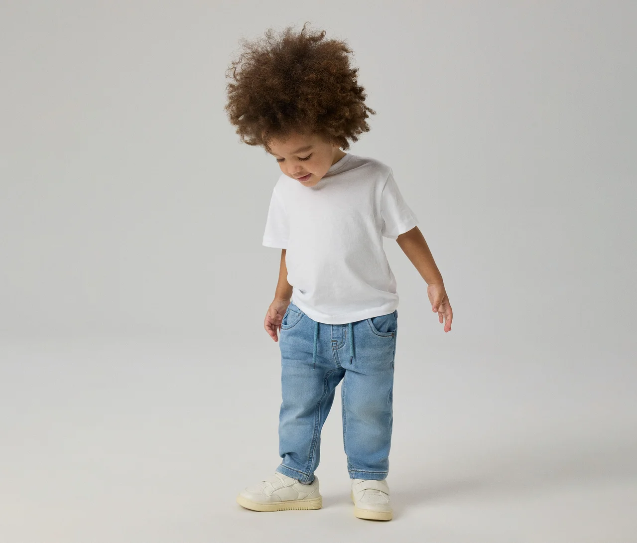 Göndör hajú kisgyermek fehér pólóban, világoskék NAME IT Slim Sweat Jeans farmerben és világos tornacipőben, lefelé néz.