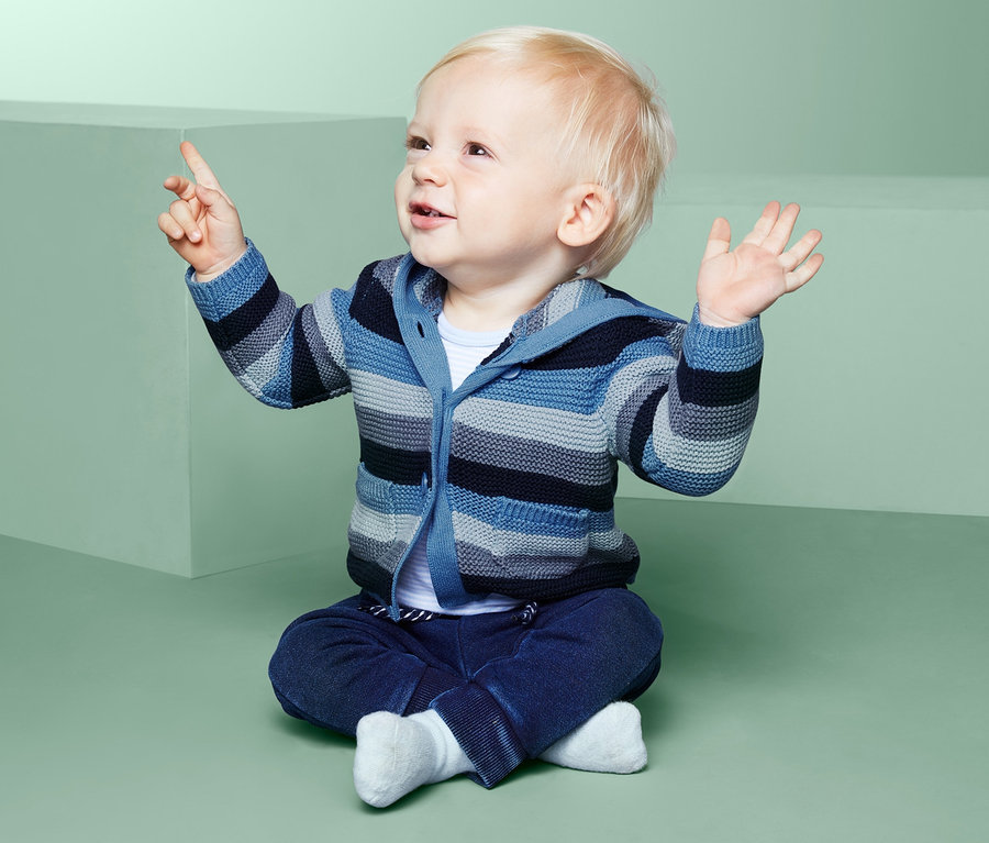 Egy világos bőrű csecsemő ül és a kötött kardigánjára mutat.