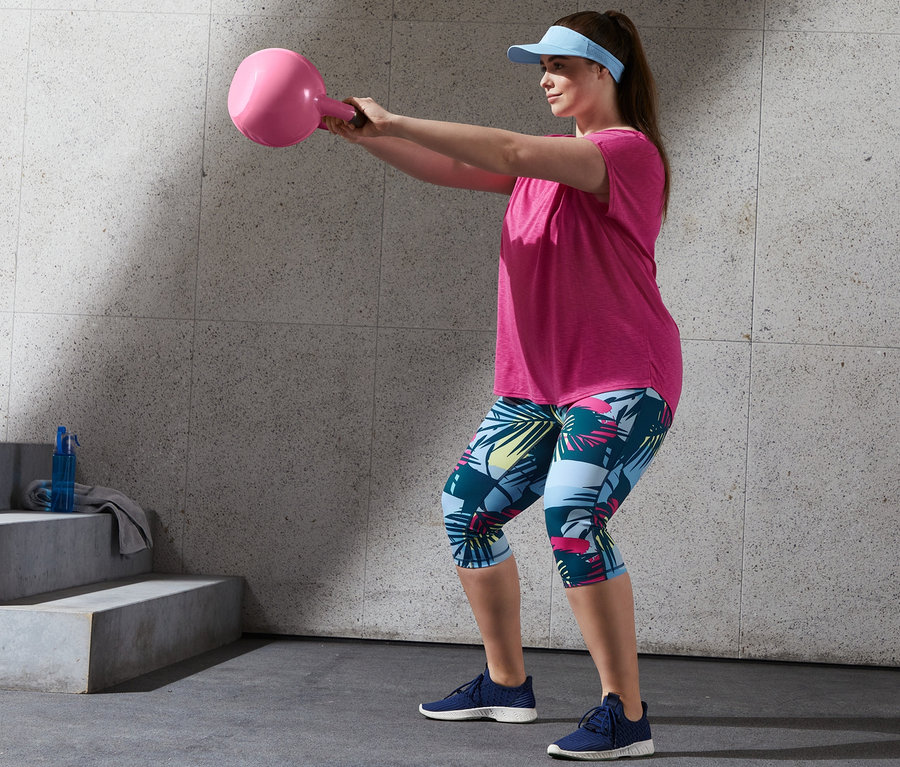 Nő rózsaszín sportfelsőben és 3/4-es sportnadrágban edz egy rózsaszín kettlebell-lel.