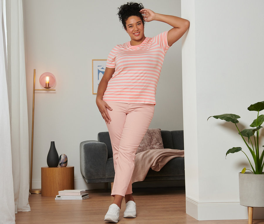 Nő pózol a falnak dőlve rózsaszín csíkos pólóban, rózsaszín slim fit farmernadrágban és velúrbőr sneaker cipőben.