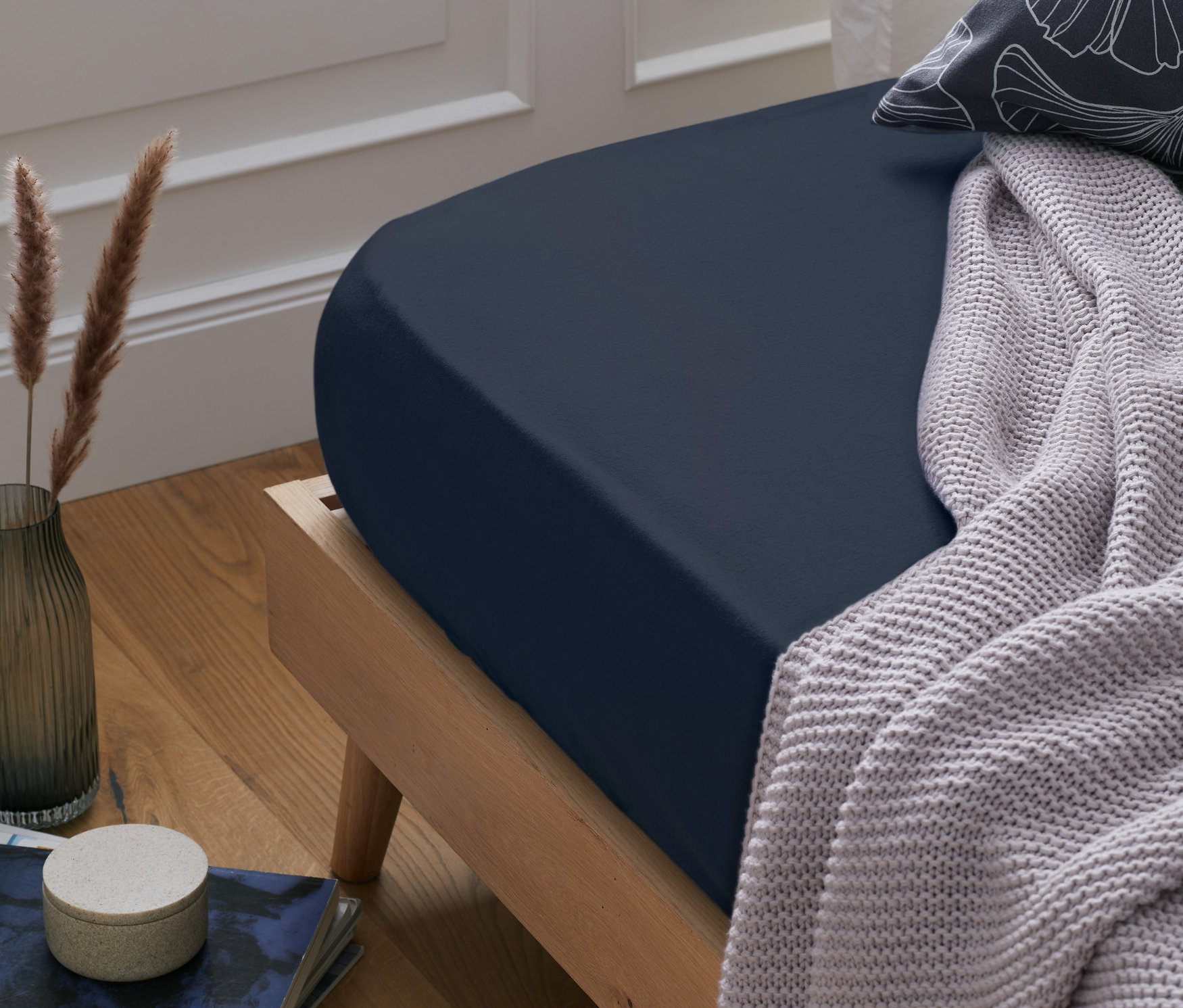 Sötétkék gumis pamutflanel lepedő egy ágyon. Mellette egy takaró és egy párna található.