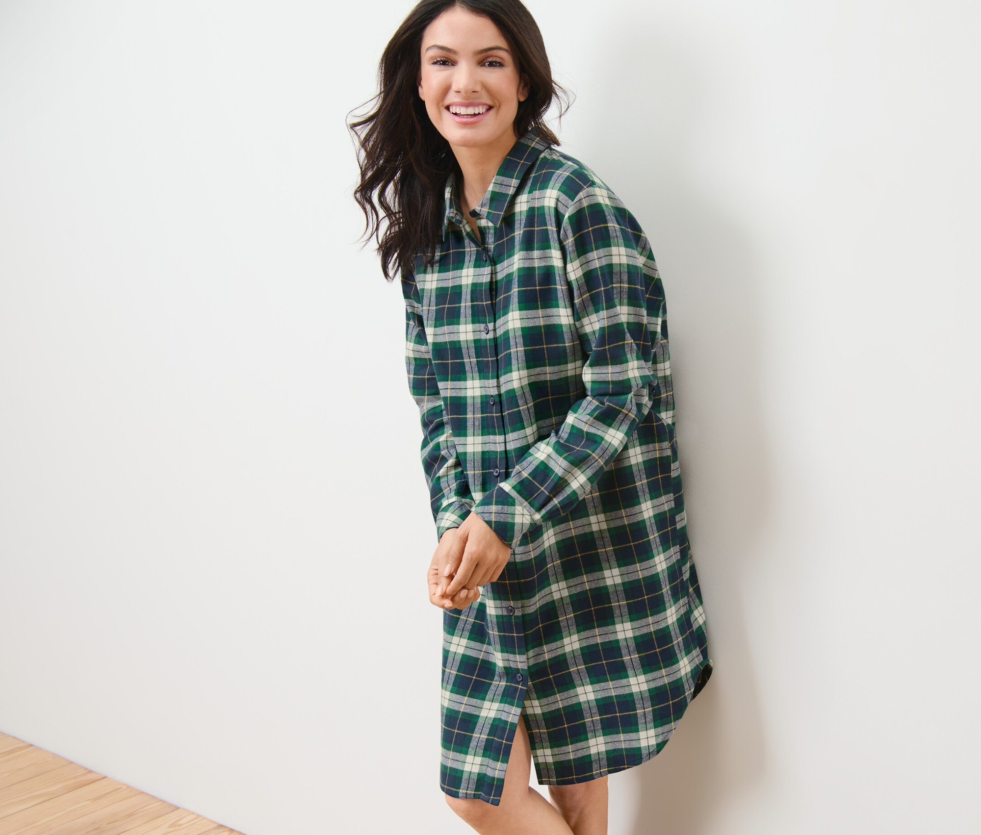 Mosolygó nő zöld pamutflanel hálóingben a falnak dőlve.